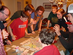 nine people huddle with eager expressions, around a dining room table with a game board at the center